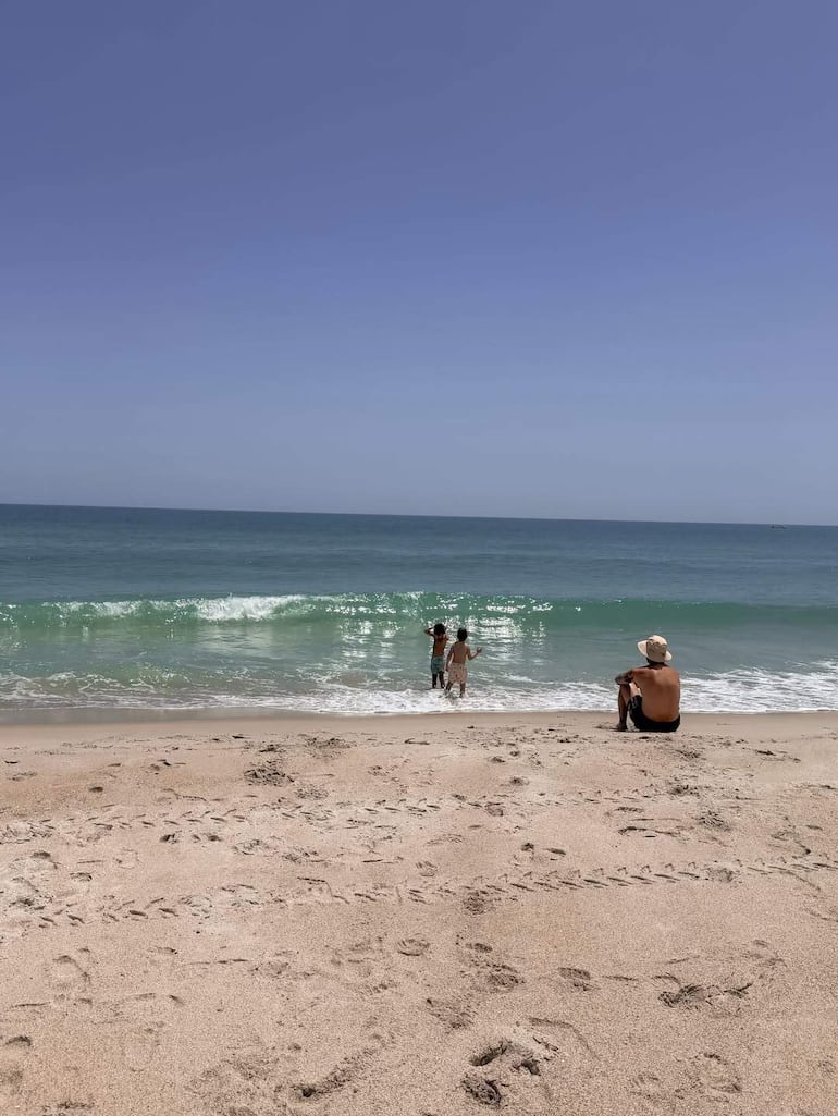 Niños jugando en el agua del mar bajo un cielo despejado, mientras un adulto en gorra blanca los observa en la arena.