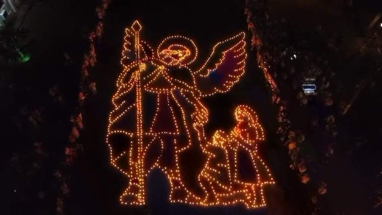 Montaje iluminado con velas formando un ángel, rodeado de personas en un ambiente festivo y exterior.