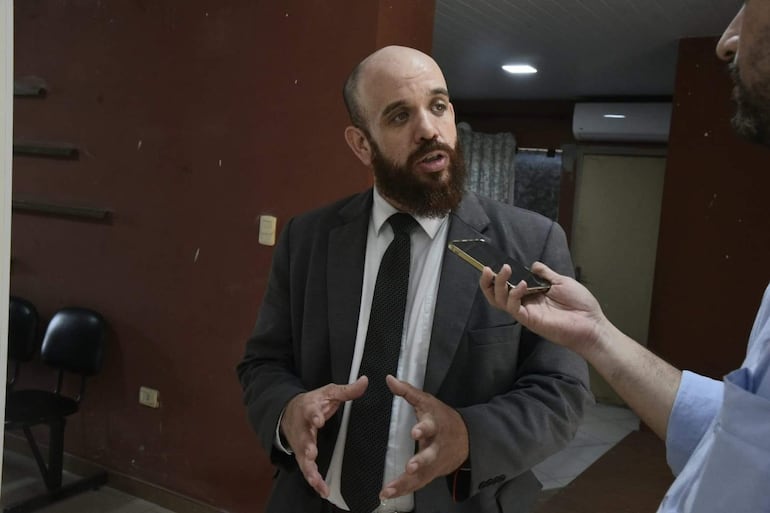 Hombre con barba y corbata gesticula mientras habla, acompañado por otro hombre que graba la conversación con un móvil.