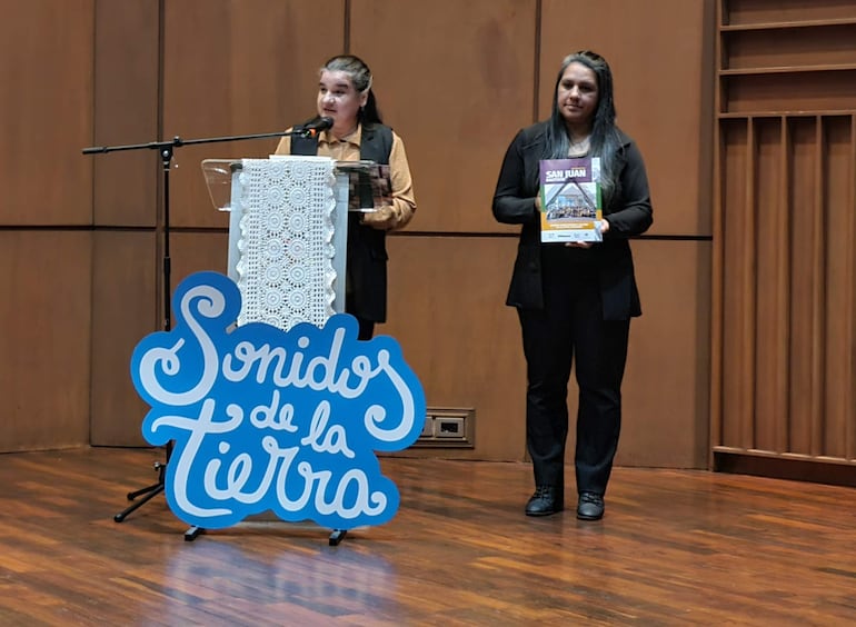 Rosa Torres habla al micrófono mientras Romina Frutos sostiene un folleto titulado 'SAN JUÁN' en un ambiente marrón formal.