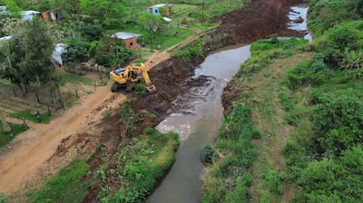 Retroexcavadora trabaja en la orilla de un arroyo en un paisaje rural, rodeada de vegetación y casas dispersas.