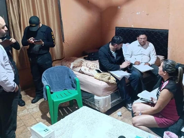 Tres hombres, uno en uniforme policial, toman notas en una habitación modesta. Mujer observa con un teléfono en la mano.
