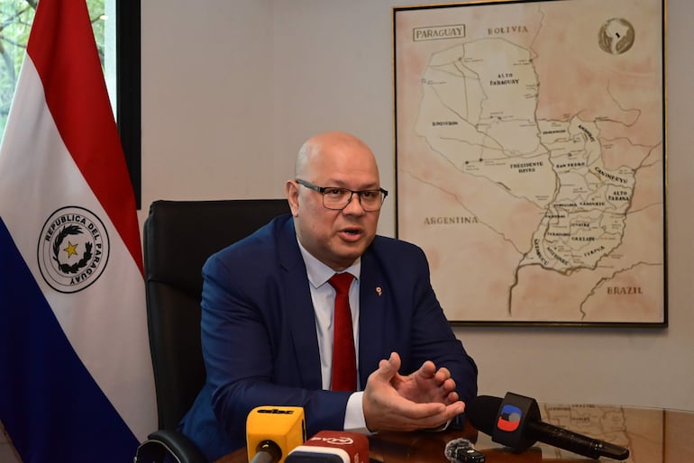 Oscar Lovera, de traje azul y corbata roja, habla en conferencia, acompañado por la bandera de Paraguay y un mapa en la pared.