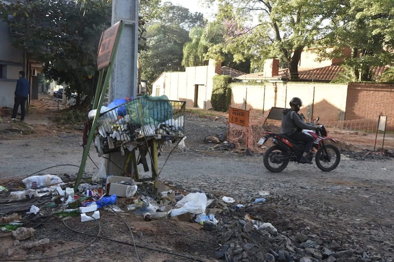 Hombre de espaldas con chaqueta azul junto a un basurero lleno de desechos en una calle residencial.
