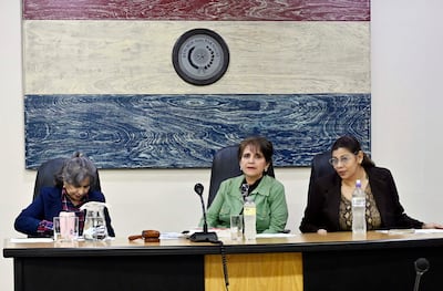 Tres mujeres sentadas en mesa: una con blazer azul, otra en chaqueta verde, y la última con blusa negra y gafas, en ambiente formal.