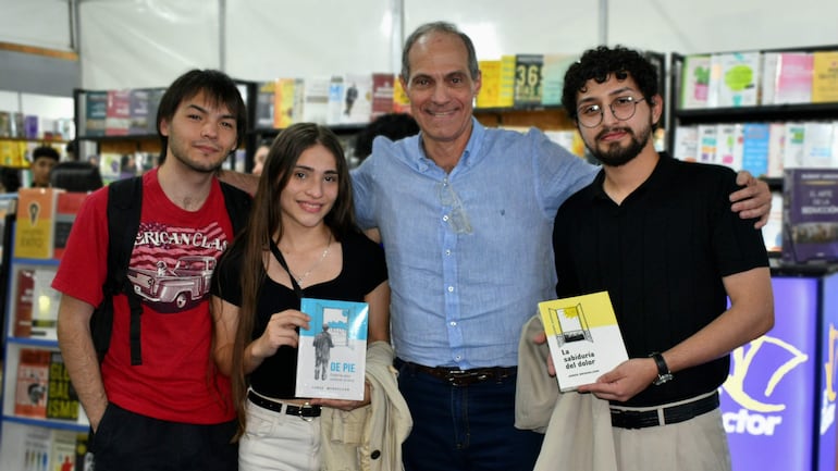 Jorge Mendelzon sonríe junto a una mujer con libro azul y un hombre con gafas, en un ambiente festivo de feria del libro.