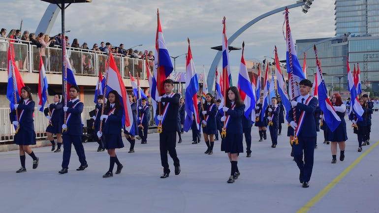 Jóvenes en uniformes oscuros marchan sosteniendo banderas de Paraguay, rodeados de una multitud en un ambiente festivo.