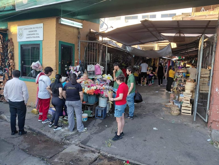 Mujer mayor rodeada de flores mientras jóvenes examinan ramos en un concurrido mercado de Asunción.