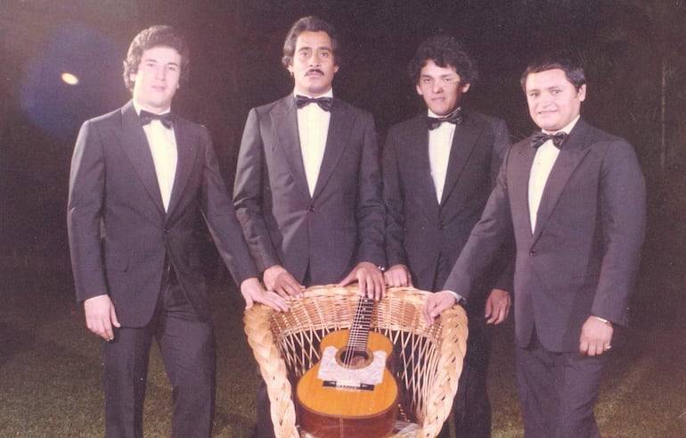 Cuatro hombres en trajes oscuros y corbatas de moño posan, con uno sosteniendo una guitarra en una canasta de mimbre.