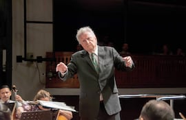 Luis Szarán de pie, gesticulando con pasión frente a músicos en un auditorio iluminado durante un concierto.