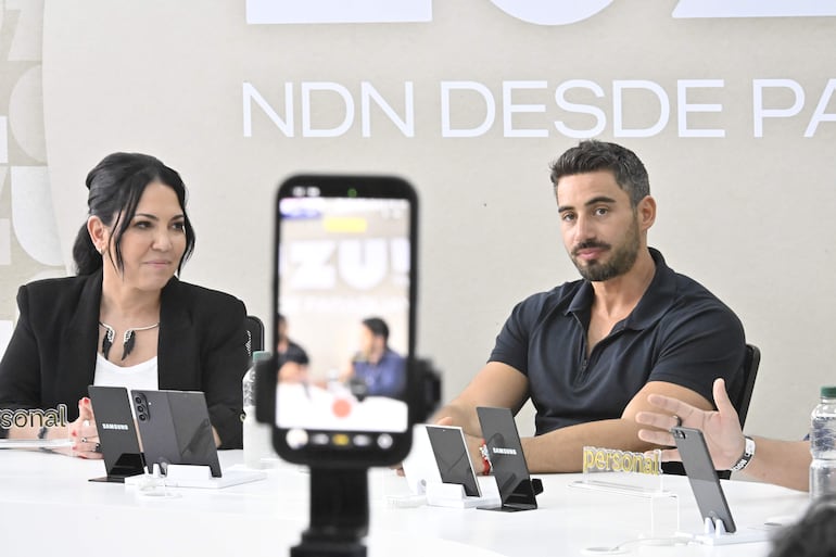 Mujer de cabello largo con chaqueta negra y blusa blanca, hombre con barba y camiseta negra en mesa blanca con teléfonos Samsung.