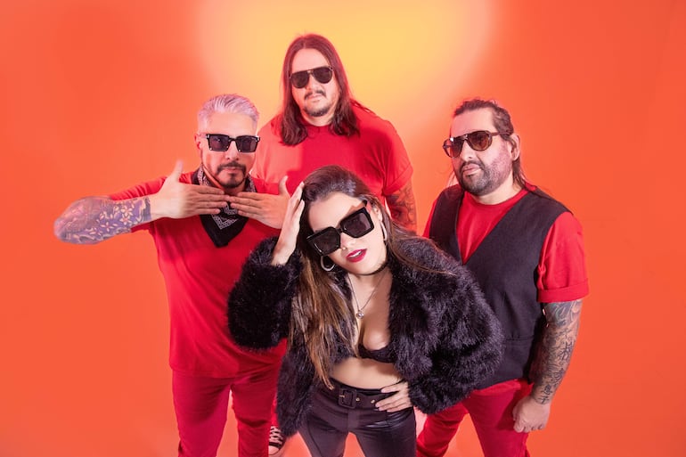 Cuatro miembros de Cielo Ámbar en primer plano, todos con gafas de sol, fondo naranja. Mujer en el centro con cabello largo y ropa negra.