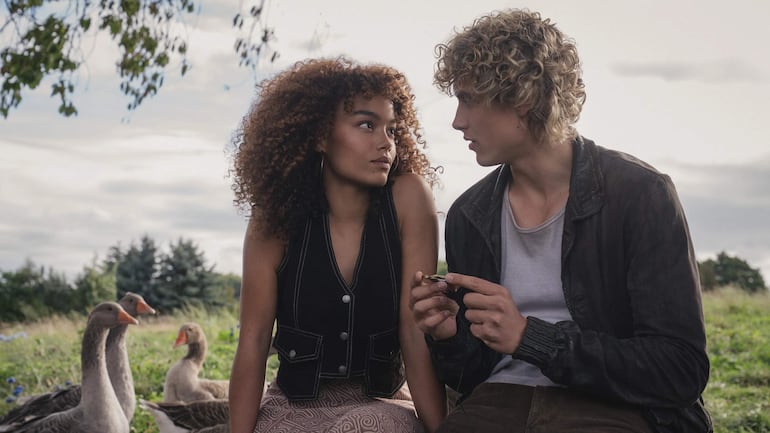 Mujer de cabello rizado y chaleco negro conversa con hombre de cabello rizado claro, rodeados de vegetación y patos en un día nublado.