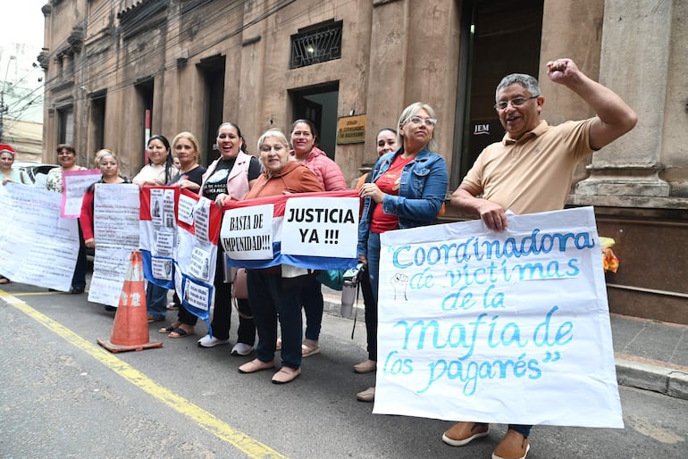 Ocho manifestantes, en su mayoría mujeres, sostienen pancartas con mensajes de protesta frente a un edificio, algunas sonríen.