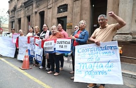 Ocho manifestantes, en su mayoría mujeres, sostienen pancartas con mensajes de protesta frente a un edificio, algunas sonríen.