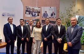 Grupo de siete personas, formalmente vestidas, sonríen en una celebración con fondo de fotografías históricas.