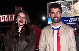 Mujer de chaqueta oscura sonríe junto a hombre en camiseta rayada y chaqueta clara, en un ambiente festivo.