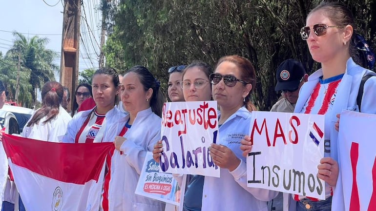 Seis bioquímicas en batas blancas sostienen carteles de protesta y una bandera de Paraguay en la manifestación.