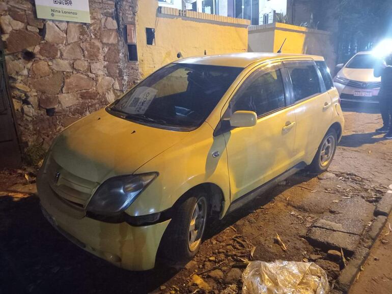 Vehículo amarillo dañado abandonado en la calle, rodeado de un entorno urbano nocturno.