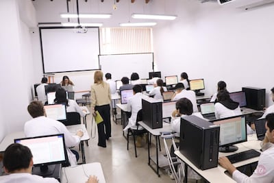 Mujer instructora de pie frente a estudiantes enfocados en computadoras, todos con batas blancas en aula iluminada.