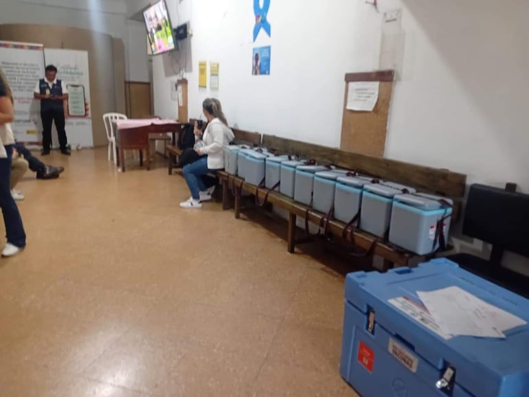 Mujer con chaqueta clara sentada en un banco, sosteniendo un teléfono móvil, mientras otras personas están presentes en un ambiente electoral.