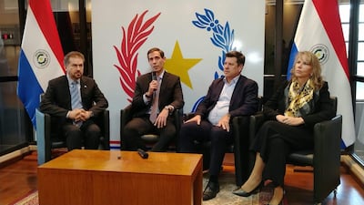 Marco Riquelme en el centro con micrófono, acompañado de Javier Giménez, Luis Castiglioni y Liz Cramer, en evento formal con banderas de Paraguay.