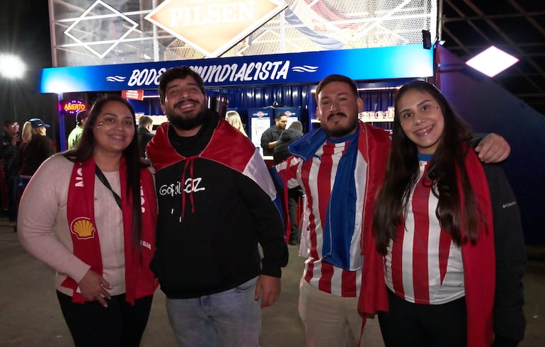 Cuatro jóvenes sonrientes en Casa Albirroja, dos en el centro con chaquetas y bufandas en los colores nacionales.