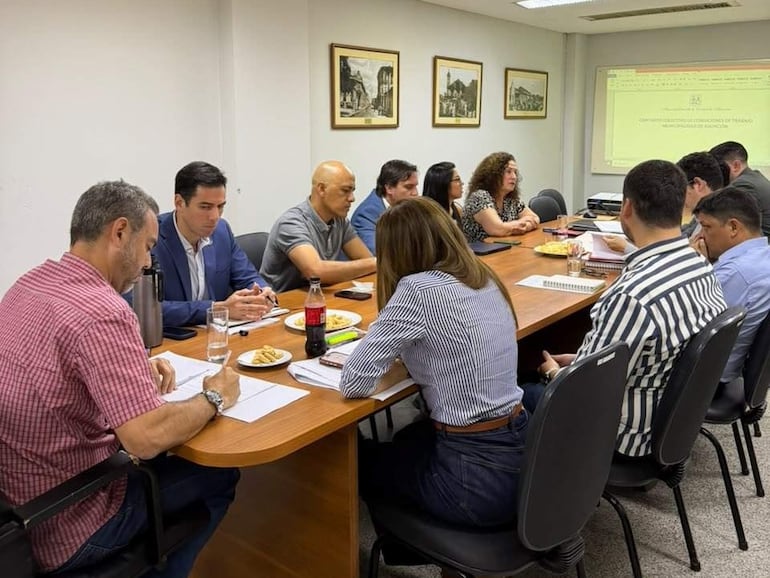 Hombres y mujeres en reunión, uno en traje azul habla mientras los demás toman notas y observan concentrados.