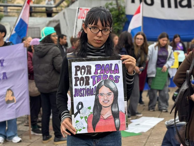 Mujer con cartel 'POR PAOLA POR TODAS JUSTICIA!' en manifestación, rodeada de personas con protestas.