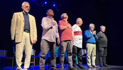 Seis hombres de pie en un escenario, vistiendo abrigos y ropa formal, participando en un acto ceremonial con gestos de respeto.