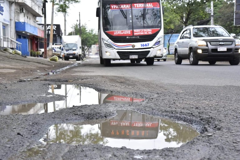 Coches y un bus en calle de Asunción, con bache visible lleno de agua, reflejando edificios y señalización.