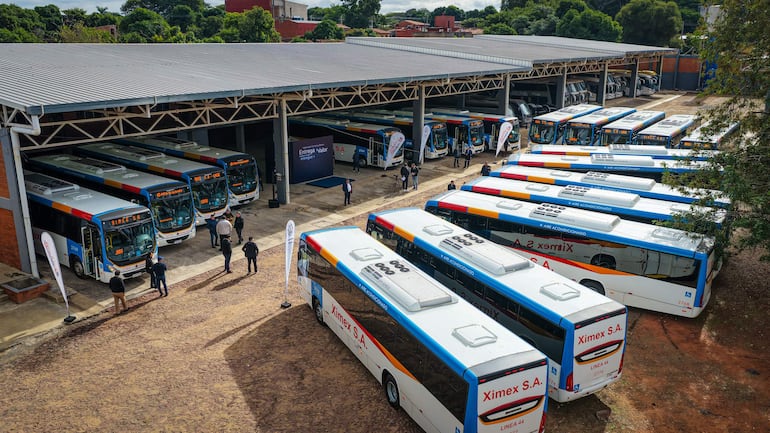 Varias personas observan autobuses de Ximex S.A., bajo un cielo nublado en un garaje amplio.