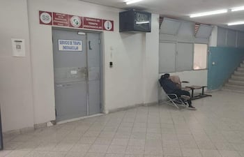 Persona sentada con abrigo oscuro en sala clínica, esperando con ambiente hospitalario y aviso de alta radiación en la pared.