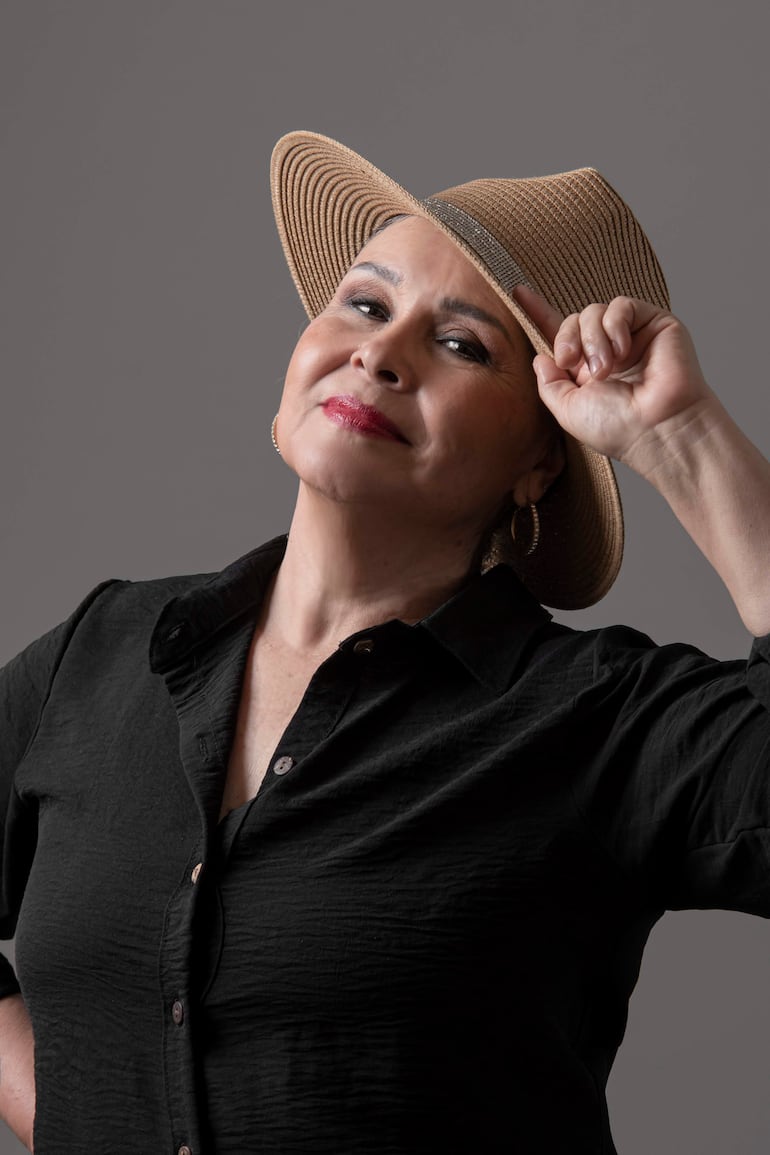 Lizza Bogado sonriente, con sombrero de ala ancha y blusa negra, levanta su sombrero con la mano en un fondo gris.