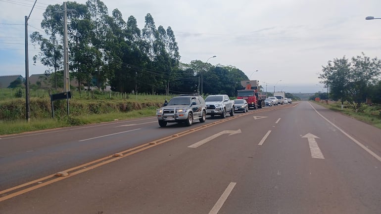 Fila de vehículos en una carretera rural, rodeada de terreno verde y árboles, sin personas visibles en la escena.