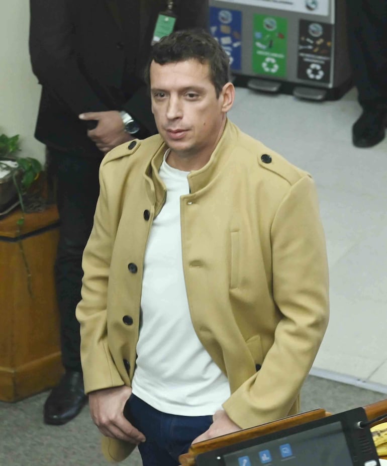 Juan Carlos Roman Galaverna con abrigo claro y camiseta blanca, en postura seria en la Cámara del Senado, junto a un guardia de negro.