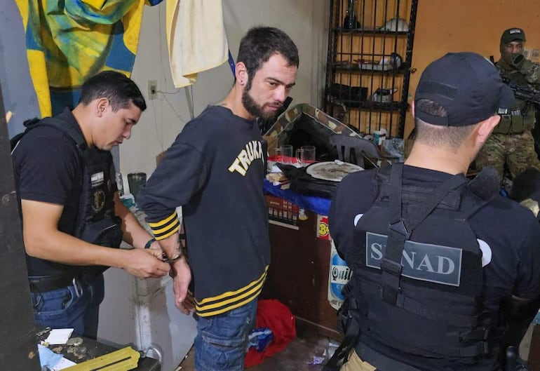 Hombre con barba y camiseta negra es esposado por agentes de la ley en un ambiente desordenado con objetos diversos.