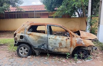 Automóvil quemado y oxidado en terreno descuidado, rodeado de vegetación, sin personas presentes.