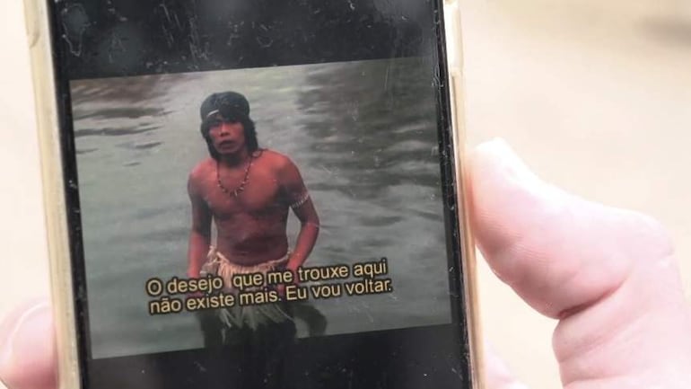 Hombre de piel morena sin camiseta sostiene un teléfono mostrando un video en un entorno acuático, con expresión tranquila.