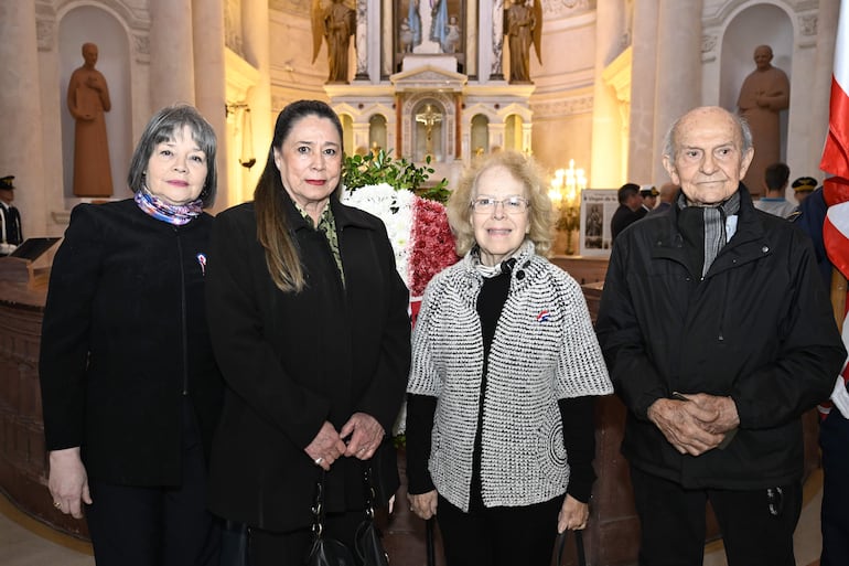Cuatro adultos, uno con cabello gris y chaqueta oscura, otro con chaqueta negra, un tercero con abrigo de rayas y anteojos, y un hombre mayor con chaqueta negra, posando en un altar iluminado.