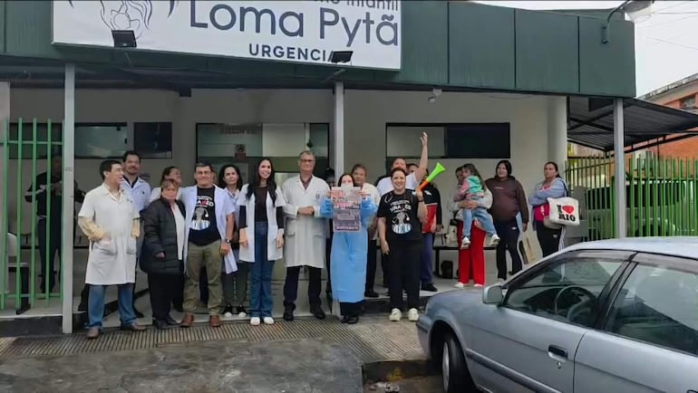 Médico con bata blanca sostiene un cartel, rodeado de colegas. Una mujer levanta el brazo, sosteniendo un niño. Ambiente urbano en Loma Pytã.