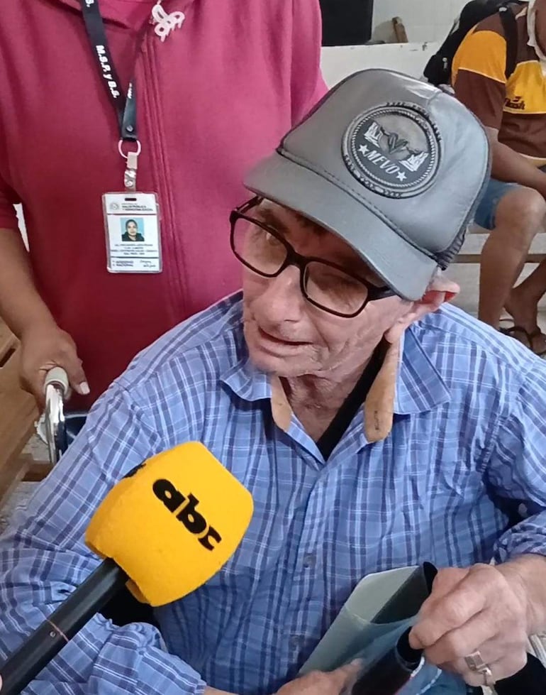 Hombre mayor sentado con gorra gris y gafas, siendo entrevistado, mientras una mujer con abrigo rojo sostiene documentos a su lado.
