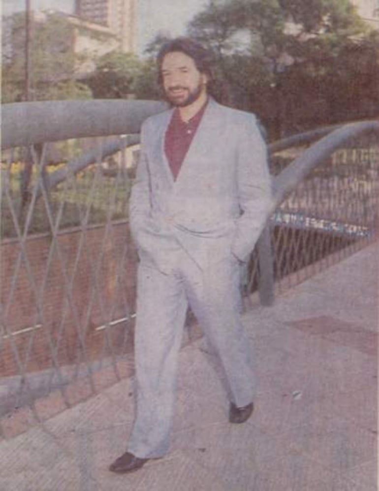 Marco Antonio Solís camina con traje claro y camiseta burdeos, con expresión feliz y manos en los bolsillos, rodeado de un entorno urbano.