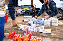 Dos agentes de la PRF agachados, manipulando paquetes de cigarrillos en un entorno exterior, con camioneta visible al fondo.