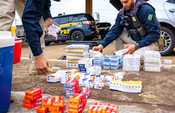 Dos agentes de la PRF agachados, manipulando paquetes de cigarrillos en un entorno exterior, con camioneta visible al fondo.