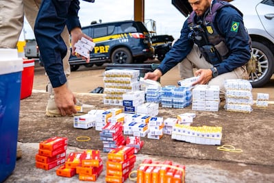 Dos agentes de la PRF agachados, manipulando paquetes de cigarrillos en un entorno exterior, con camioneta visible al fondo.