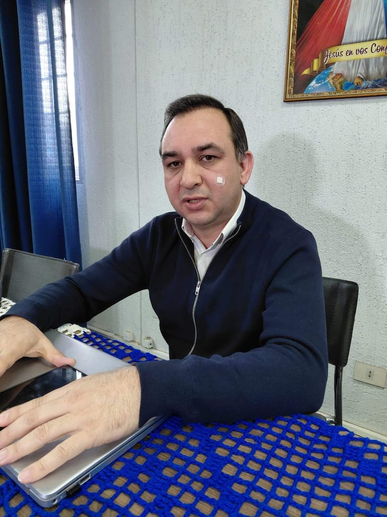 Hombre sentado con suéter oscuro y camisa blanca, vendaje en el rostro, rodeado de computadora portátil y teléfono en mesa con mantel azul.