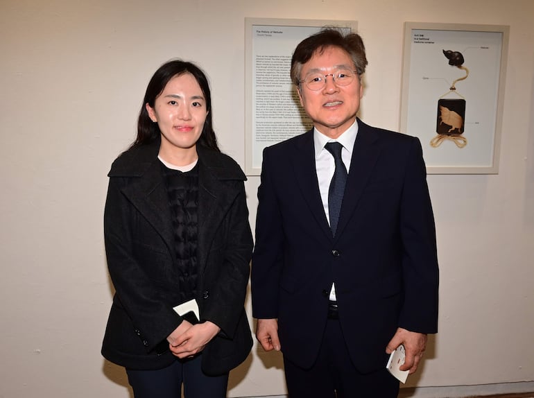 Yuushi Tanida con chaqueta oscura y mujer con cabello largo, sosteniendo objetos en un ambiente de exposición, sonriendo.