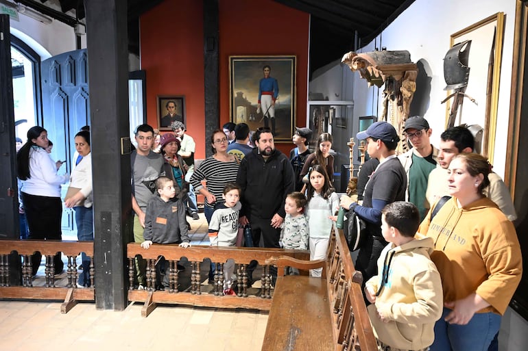 Grupo de personas en sala de museo, con niños curiosos y adulto explicando, en ambiente decorado con historias y artefactos.