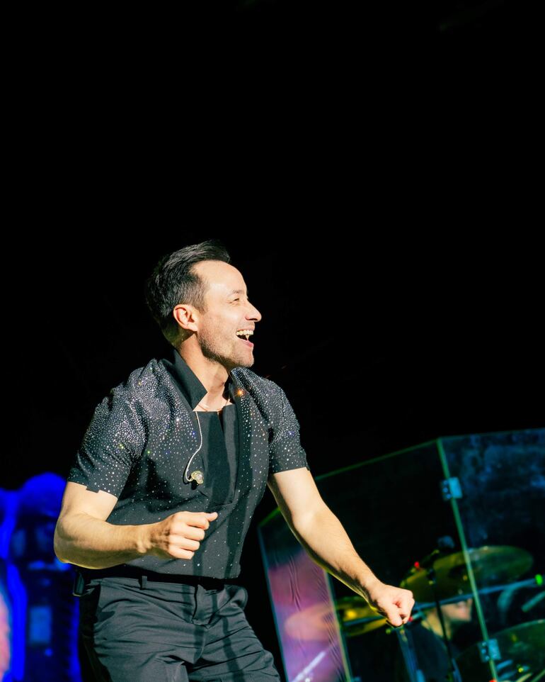 Hombre sonriendo y bailando en el escenario, vestido de oscuro con detalles brillantes, energía contagiosa en un entorno musical.